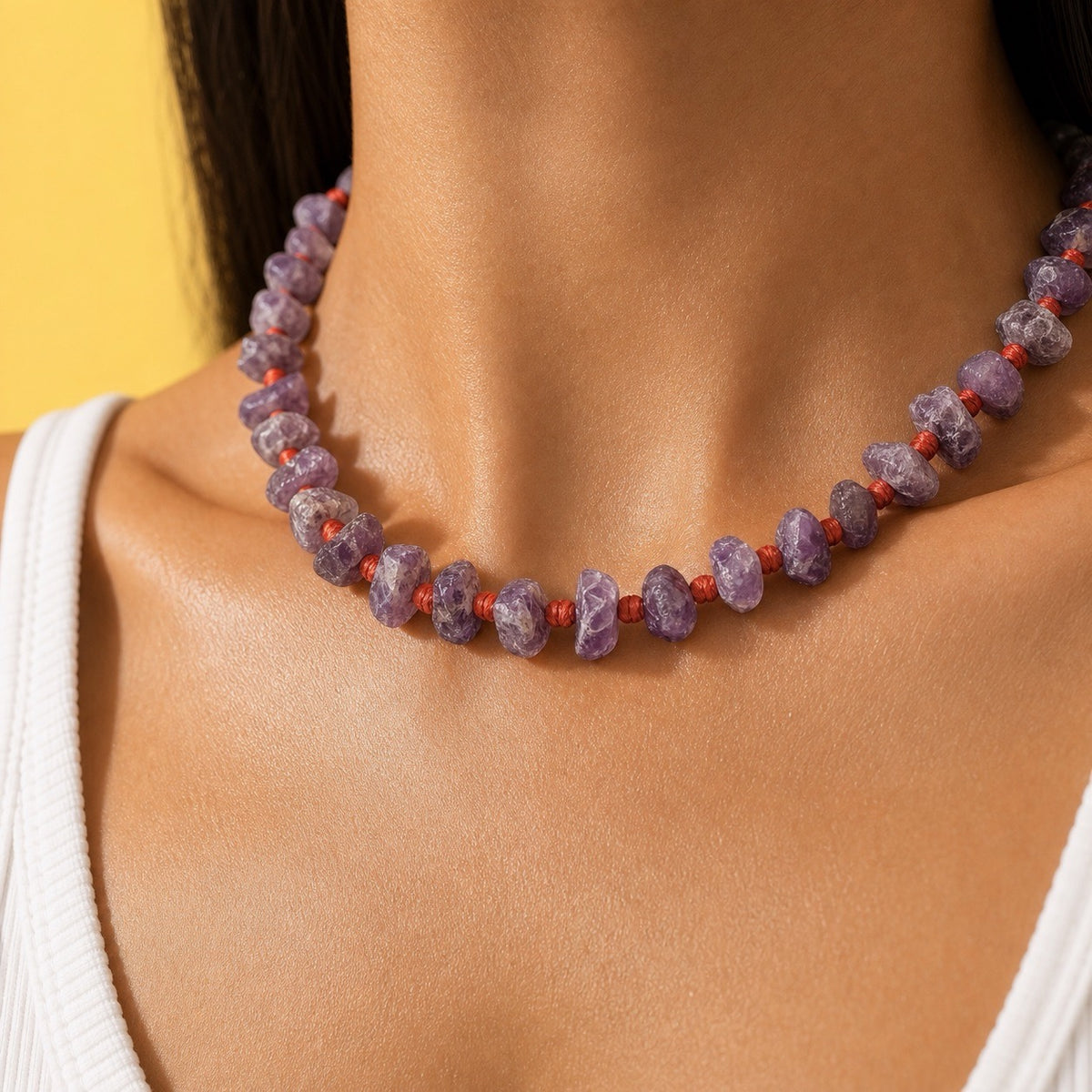 A close-up of a person's neck and upper chest wearing a beaded necklace made of irregular purple stones separated by small red cord knots, against a yellow background.
