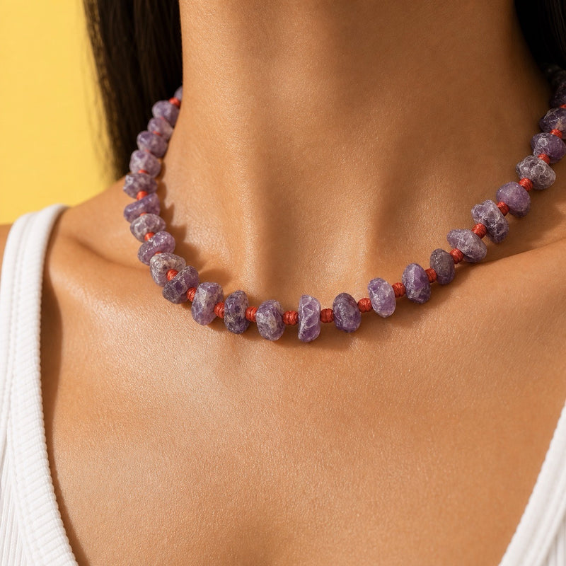 A close-up of a person's neck and upper chest wearing a beaded necklace made of irregular purple stones separated by small red cord knots, against a yellow background.