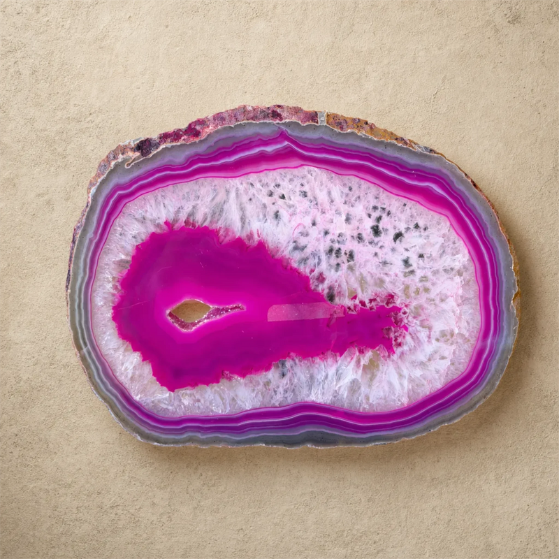 A polished slice of agate or quartz geode with vivid magenta pink, purple, white, and gray banded layers, photographed from above on a textured beige surface.