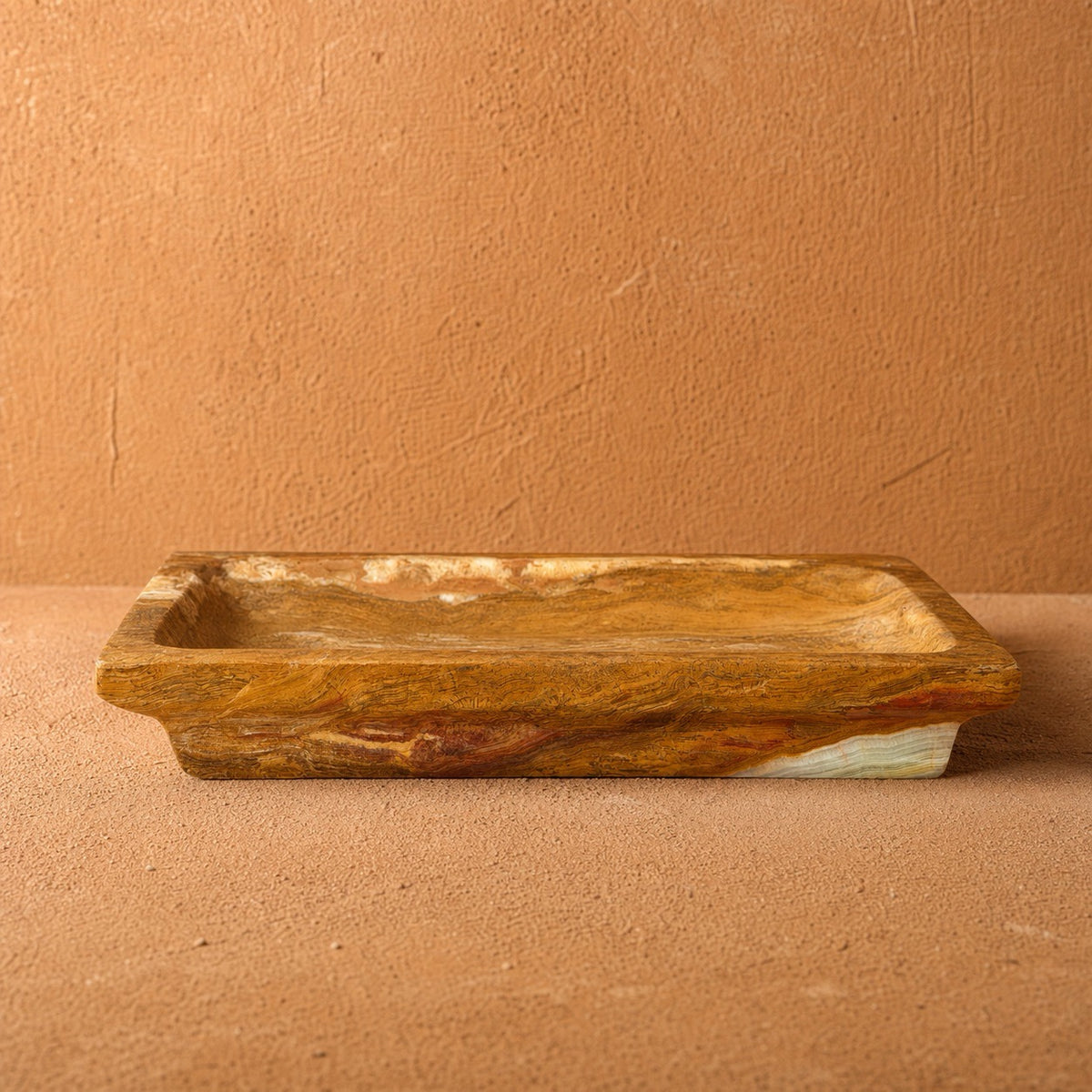 A rectangular stone tray with a rustic, hand-carved texture in earthy brown, gold, and orange tones, set against a matching terracotta background.