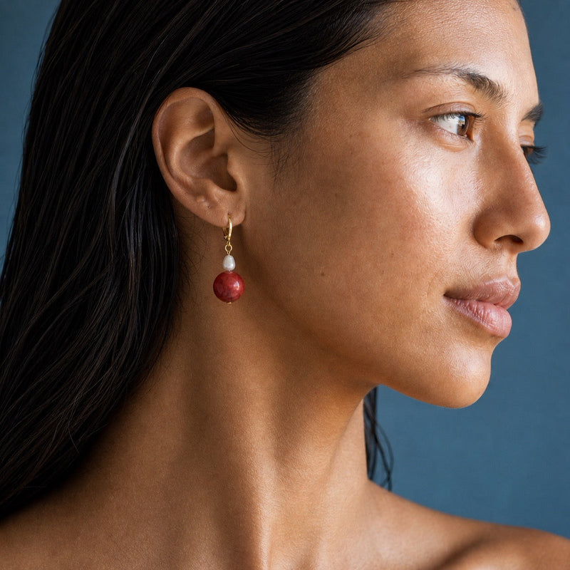 A close-up side profile of a woman wearing a single drop earring with a small white freshwater pearl and a round red coral bead suspended from a gold hook.