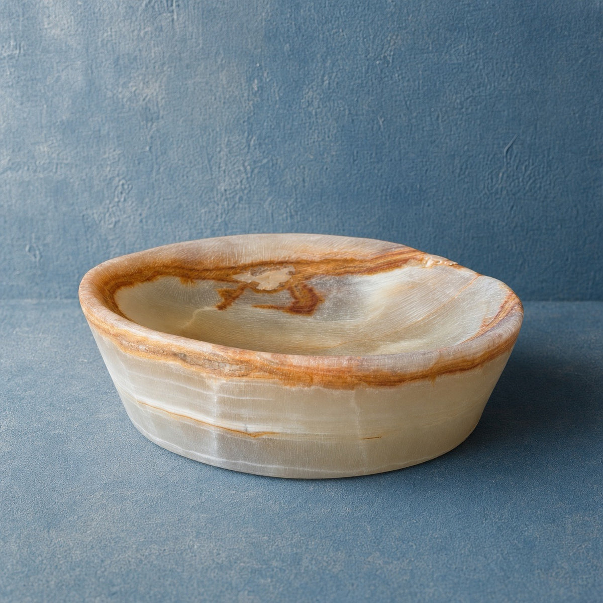 A shallow oval onyx bowl made from natural onyx with swirling bands of cream, beige, and warm brown, set against a textured blue-gray background.