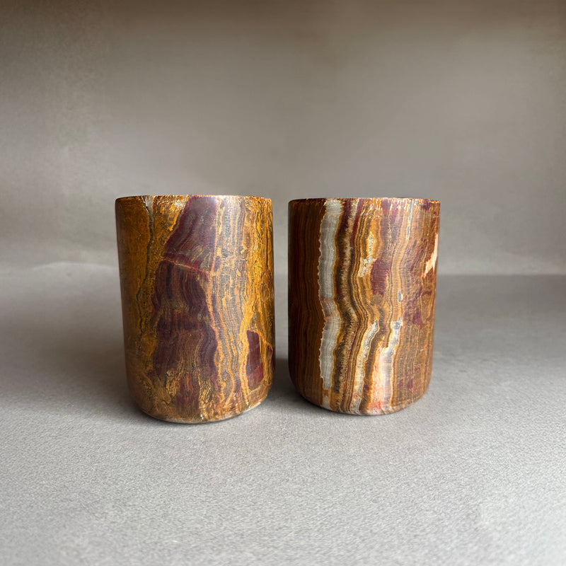 Pair of hand-carved natural onyx vessels in rust, amber, and caramel tones with cream veining — minimalist cylindrical planters or pen holders by SAÉA.