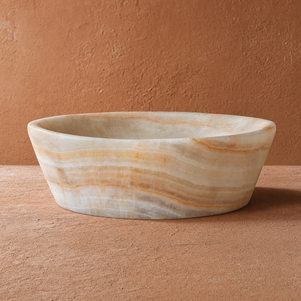A round  peach onyx stone vessel bowl with organic beige, cream, and light brown marbled veining, set against a textured terracotta background.