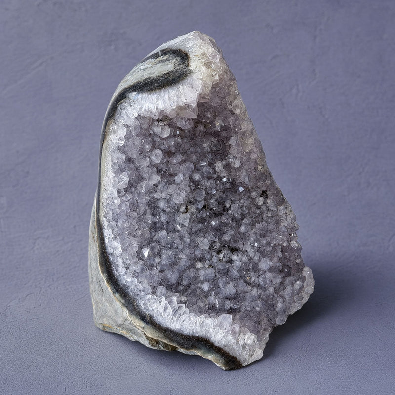 A natural crystal amethyst geode specimen with a white outer matrix and a gray-purple crystalline interior, standing upright on a textured gray background.