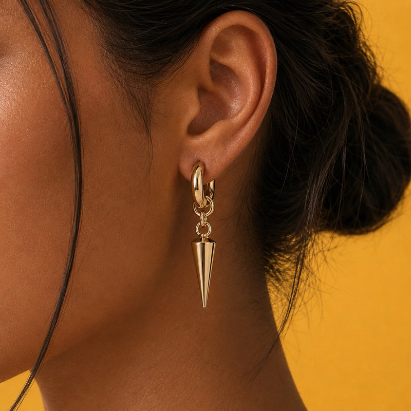 A close-up of a model's ear wearing a gold hoop earring with a dangling metallic cone charm, set against a solid yellow background.