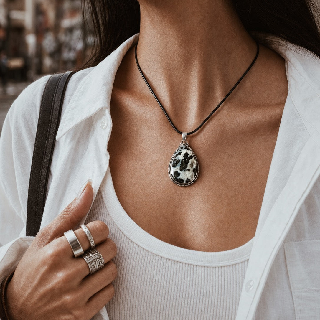 A close-up lifestyle shot of a woman wearing a teardrop-shaped pendant necklace made from a black and white marbled stone, suspended on a black cord. She is wearing a white button-down shirt over a white ribbed tank top, with rings on her fingers.