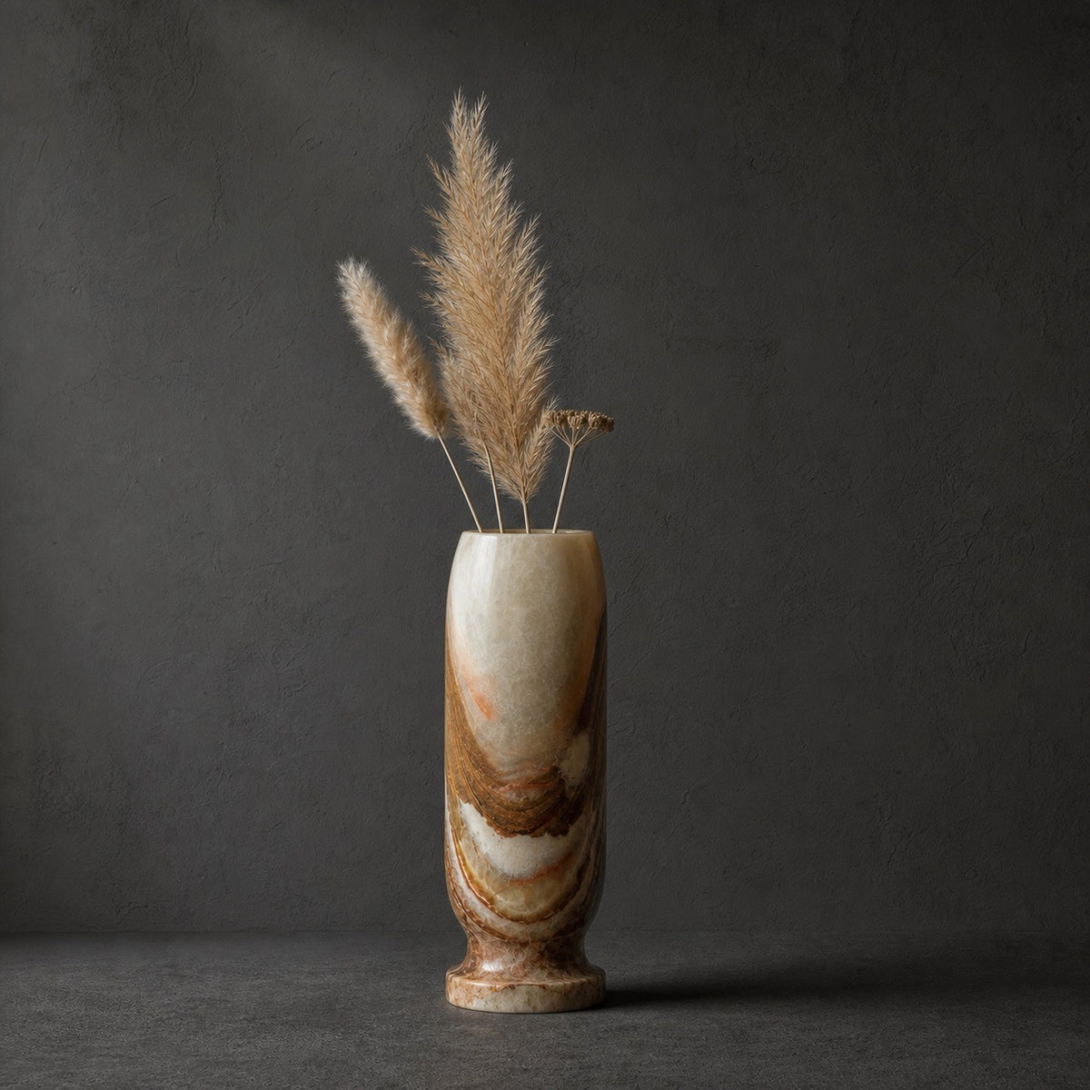 Sage and rust onyx origin vase holding dried pampas grass, its pale column banded with rust and amber toward the footed base, against a dark grey wall.