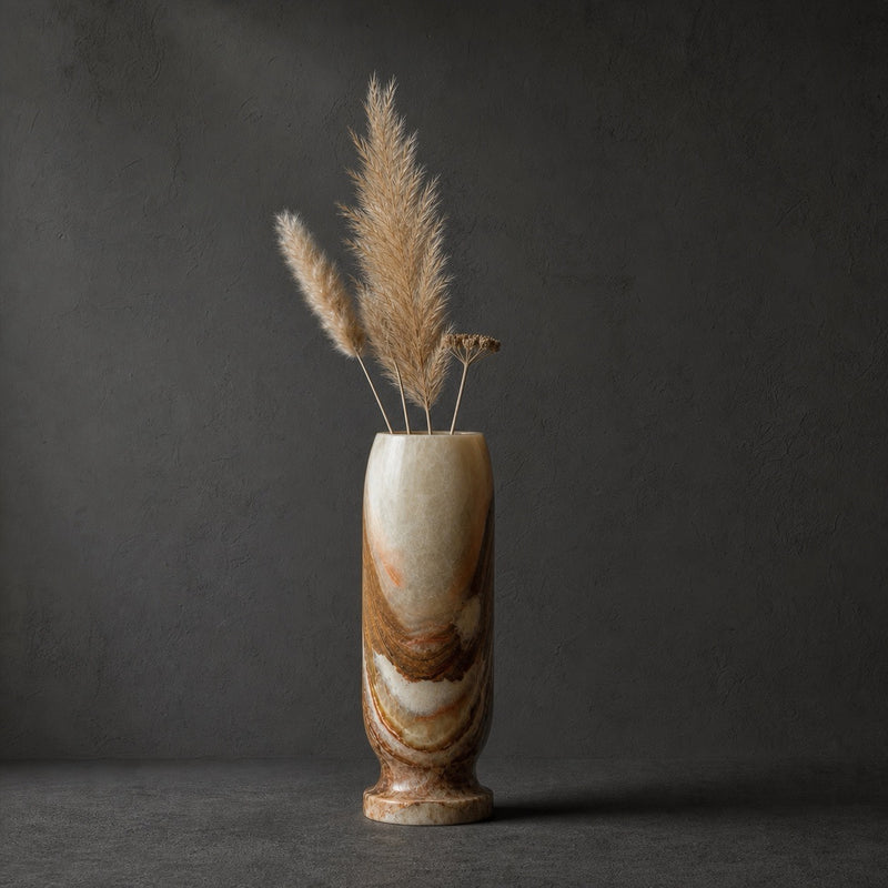 Sage and rust onyx origin vase holding dried pampas grass, its pale column banded with rust and amber toward the footed base, against a dark grey wall.