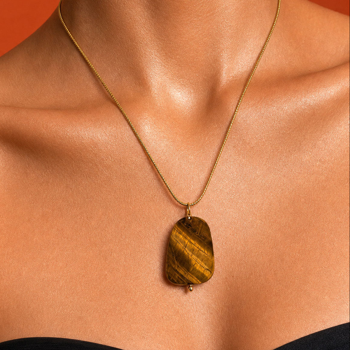 A gold chain necklace with a polished tiger's eye stone pendant, worn on a model's neck against a solid orange background.