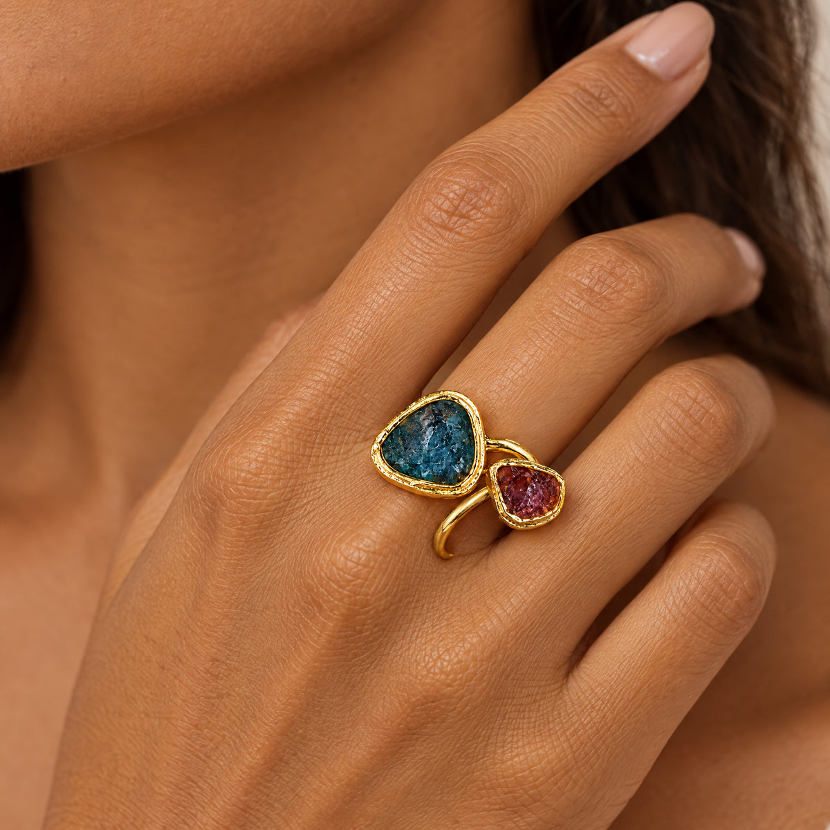 Raw apatite and ruby bypass ring worn on a hand raised to the collarbone, the teal blue apatite and rose red ruby in hammered 22k gold-filled bezels