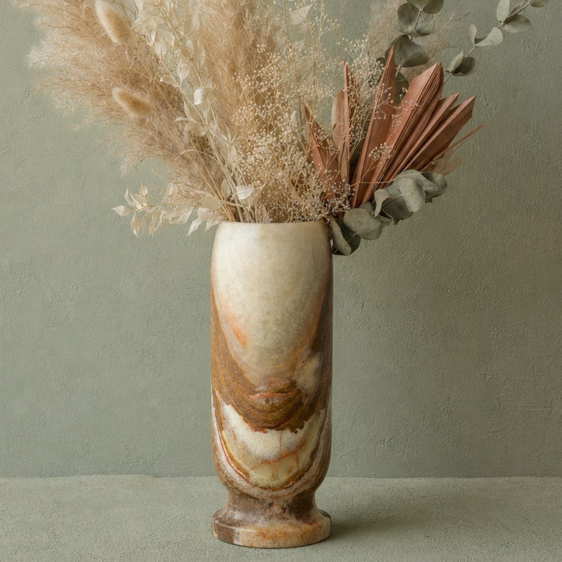 A tall cylindrical green onyx vase with a marbled beige, brown, and cream pattern, filled with dried decorative grasses and botanical stems, set against a muted sage green wall.