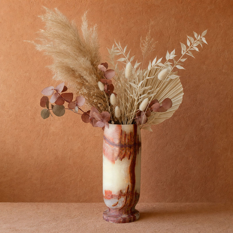 A decorative dried floral arrangement in a tall onyx marbled vase, set against a warm terracotta textured background. The arrangement features beige pampas grass, white fringed seed pods, and dusty rose eucalyptus leaves in a cream and rust marbled vase.