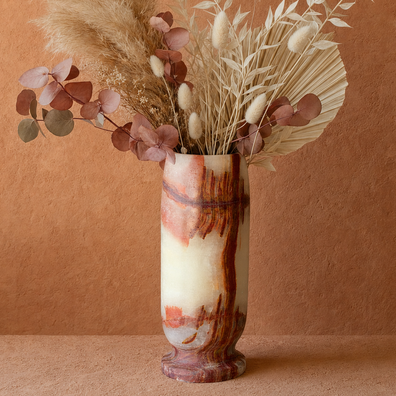 A decorative dried floral arrangement in a tall marbled vase, set against a warm terracotta textured background. The arrangement features beige pampas grass, white fringed seed pods, and dusty rose eucalyptus leaves in a cream and rust marbled vase.