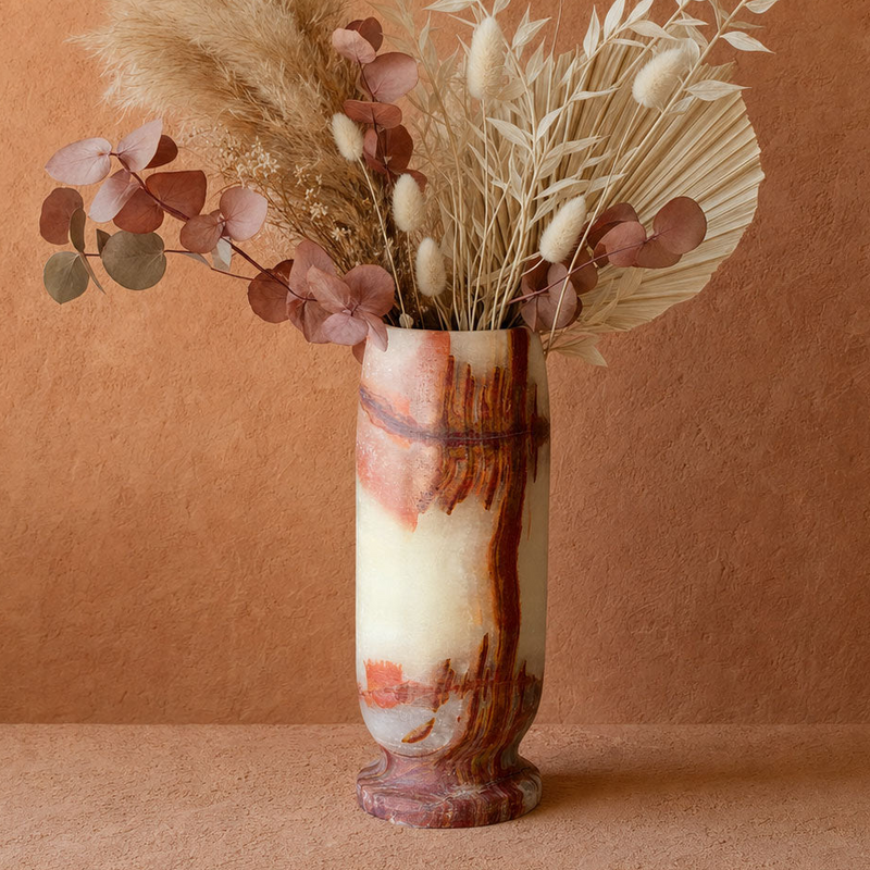 A tall cylindrical vase with a marbled cream, terracotta, and brown pattern, filled with dried pampas grass, eucalyptus leaves, and a palm frond, set against a textured terracotta background.