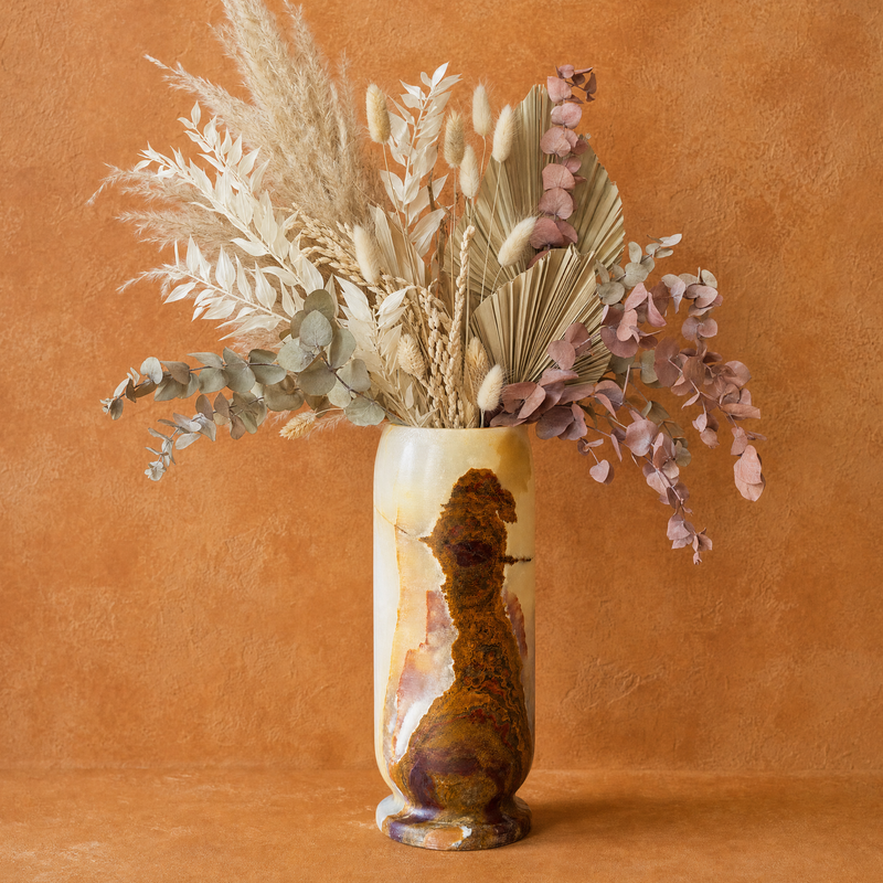 A decorative onyx vase with a glossy brown and gold abstract design, filled with a dried floral arrangement of beige pampas grass, eucalyptus, and dusty pink foliage, set against a warm textured terracotta background.