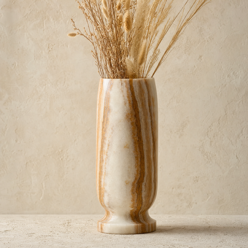 A tall cylindrical onyx vase with vertical tan and cream veining, filled with dried grasses and stems, set against a textured beige plaster wall and surface.