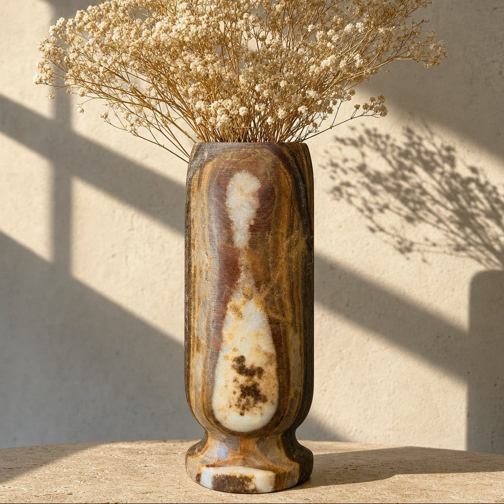 A tall cylindrical ceramic vase with a marbled brown, beige, and cream pattern, filled with dried baby's breath flowers, set on a stone surface against a textured wall with window shadow patterns.