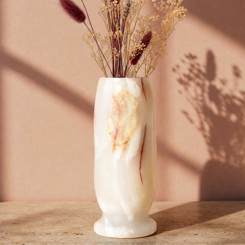 A cream-colored onyx-style vase with natural marbled gold and reddish-brown veining, filled with dried grasses and stems, set on a stone surface against a warm beige wall with soft shadows.