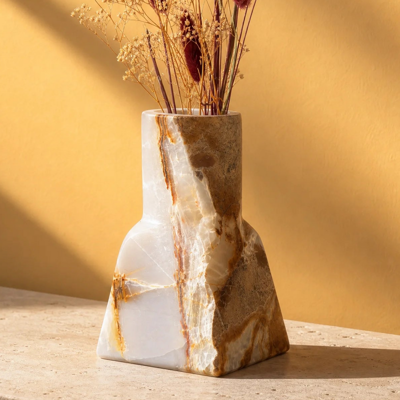 A sculptural stone vase made of white and tan marble with brown veining, holding dried grasses, set on a stone surface against a warm yellow wall with soft shadows.