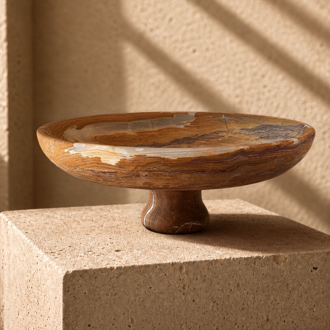 Hand-carved onyx pedestal bowl in amber and caramel with plum banding and cream patches at the rim, on a sandstone plinth in afternoon light