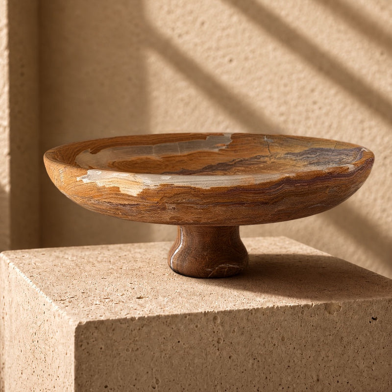 Hand-carved onyx pedestal bowl in amber and caramel with plum banding and cream patches at the rim, on a sandstone plinth in afternoon light