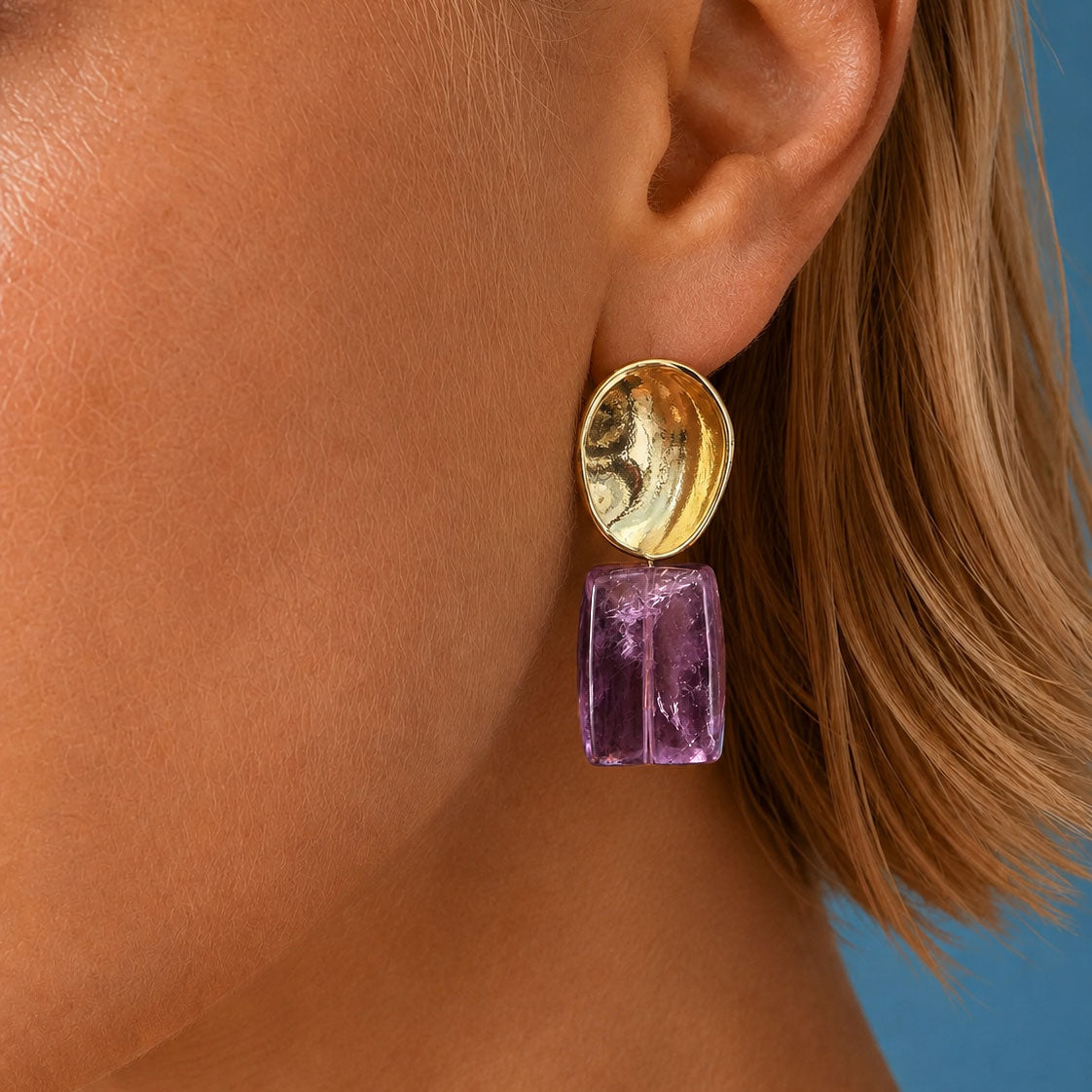 A close-up of a model's ear wearing a drop earring with a gold-toned oval top and a faceted purple rectangular stone below, set against a solid blue background.

amethyst-gold-filled-drop-earrings-worn-saea.jpg · alt: Amethyst and gold-filled drop earring worn on the ear, a concave 24k gold-filled brass disc above a raw purple amethyst tablet
