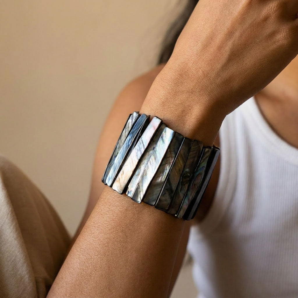 A close-up of a person's wrist wearing a wide cuff bracelet made of vertical iridescent shell segments that shift between silver, blue, and green.

Model in white tank and brown trousers wearing a wide black mother-of-pearl stretch cuff bracelet on the wrist
