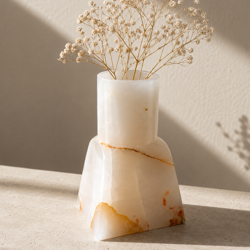 A modern two-tiered white onyx vase with organic gold and orange veining, holding dried baby's breath flowers, set on a textured beige surface with warm sunlight and shadows.