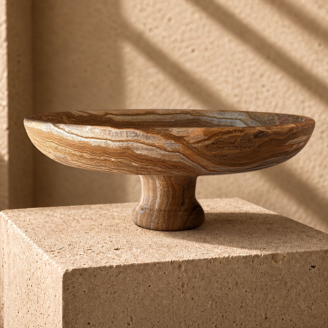 Hand-carved brown onyx pedestal bowl with walnut, caramel and silver-slate banding, set on a sandstone plinth in raking afternoon light