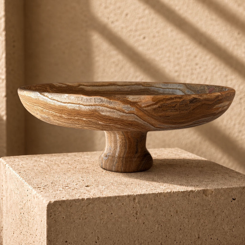 Hand-carved brown onyx pedestal bowl with walnut, caramel and silver-slate banding, set on a sandstone plinth in raking afternoon light