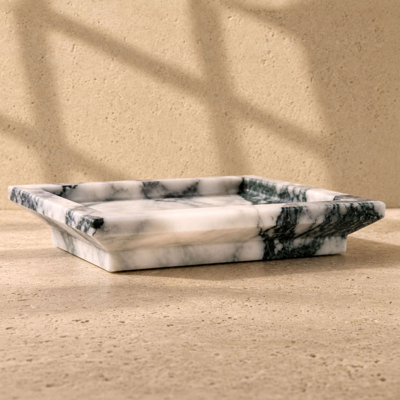A rectangular marble tray with a white base and dark green veining, set on a textured beige stone surface with soft window shadow patterns. White Calacatta Vagli marble catchall tray with deep green and charcoal veining, raised rim on a plinth base, on a travertine surface
