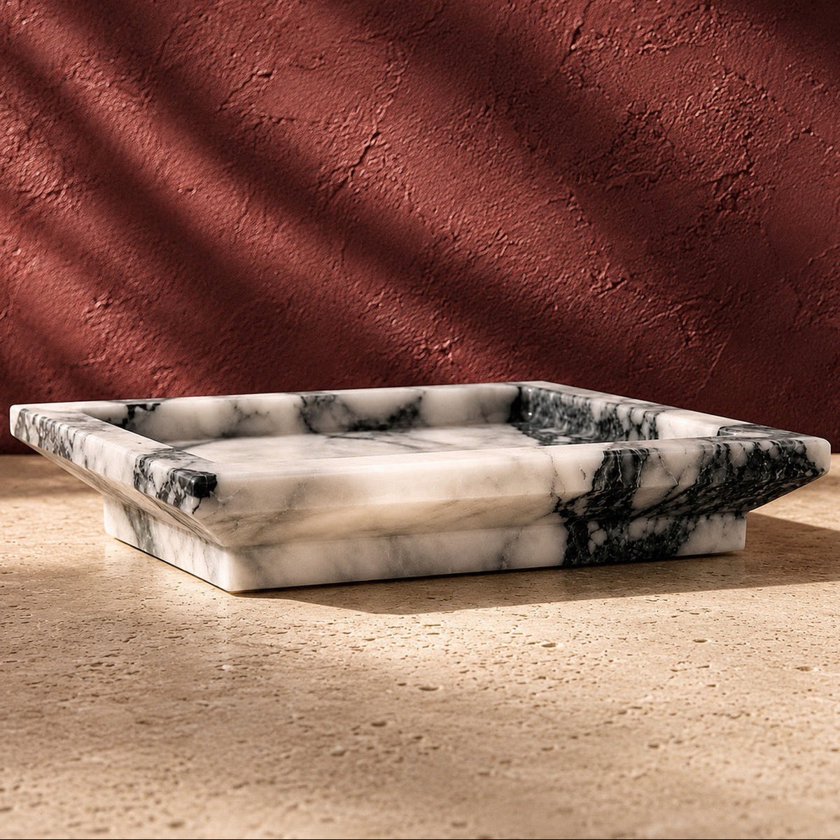 A rectangular calacatta vagli marble tray with black and gray veining, set on a textured beige stone surface against a terracotta wall with dramatic leaf shadows.