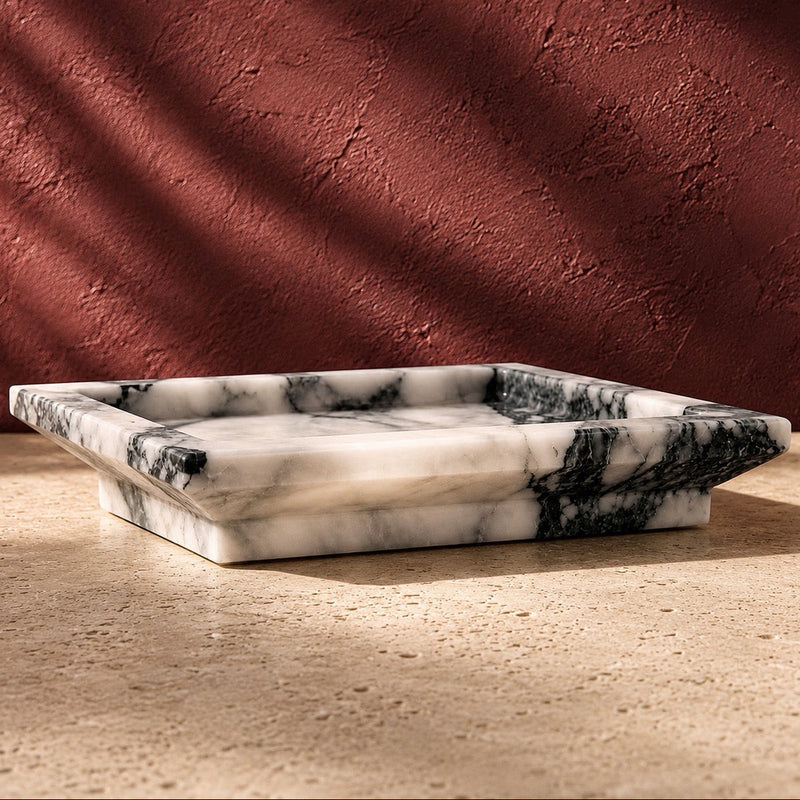 A rectangular calacatta vagli marble tray with black and gray veining, set on a textured beige stone surface against a terracotta wall with dramatic leaf shadows.
