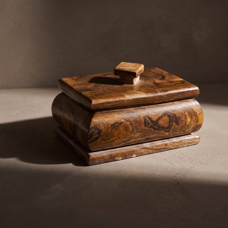 Caramel and tobacco onyx keepsake box with lid in a three-quarter front view: dark brown swirls and an oval marking on the bulged front, a stepped base and a small square stone knob, in a slant of sunlight