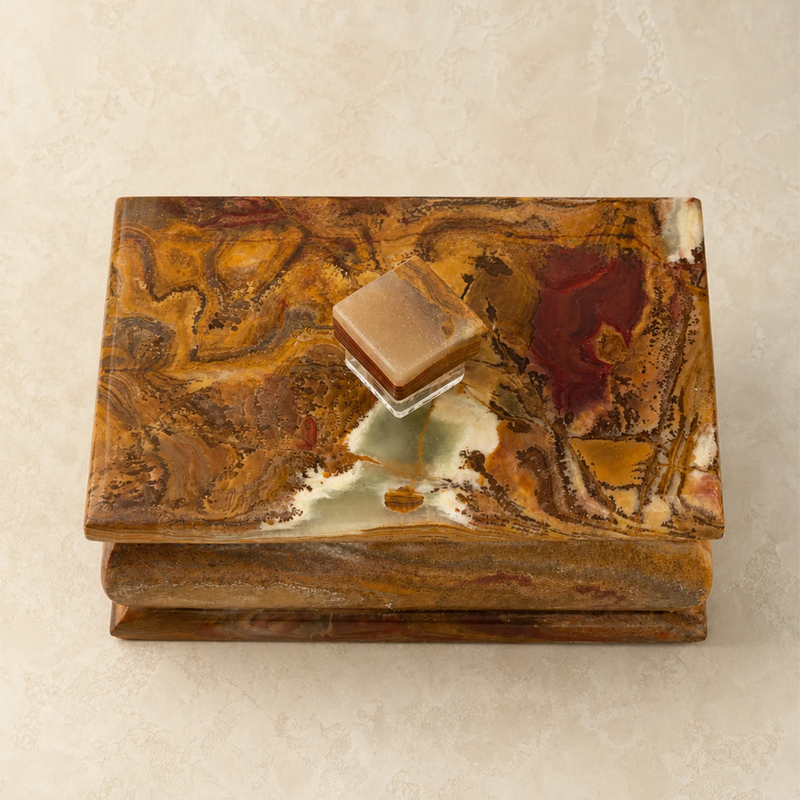 A small decorative box with a marbled stone or resin lid in earthy brown, gold, and cream tones, featuring a small square object resting on top, set against a light beige textured background.