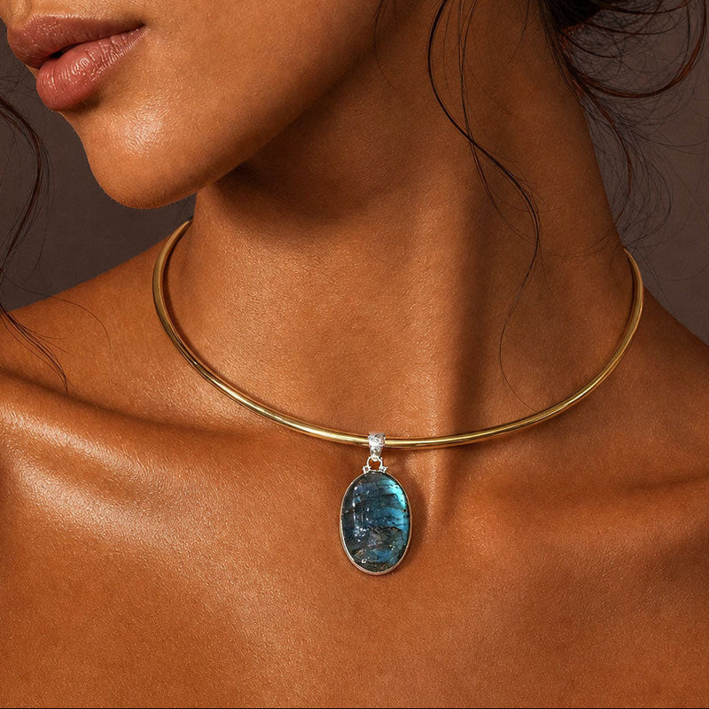 A close-up of a model wearing a delicate gold collar necklace with a dangling oval blue labradorite gemstone pendant.