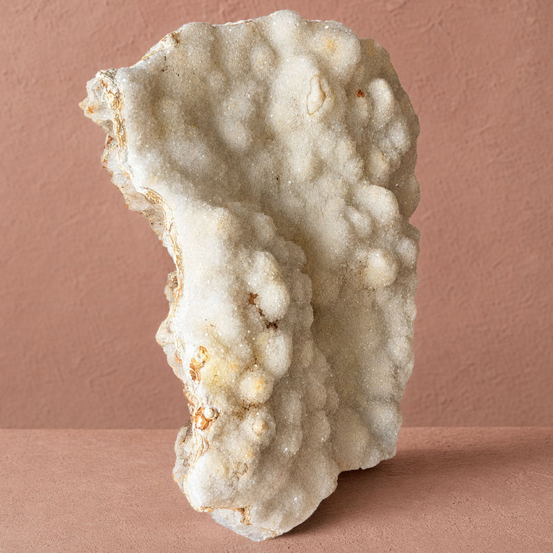 An irregular-shaped white crystalline mineral or stone specimen with a rough, bubbled texture, standing upright on a dusty pink surface against a matching pink wall.