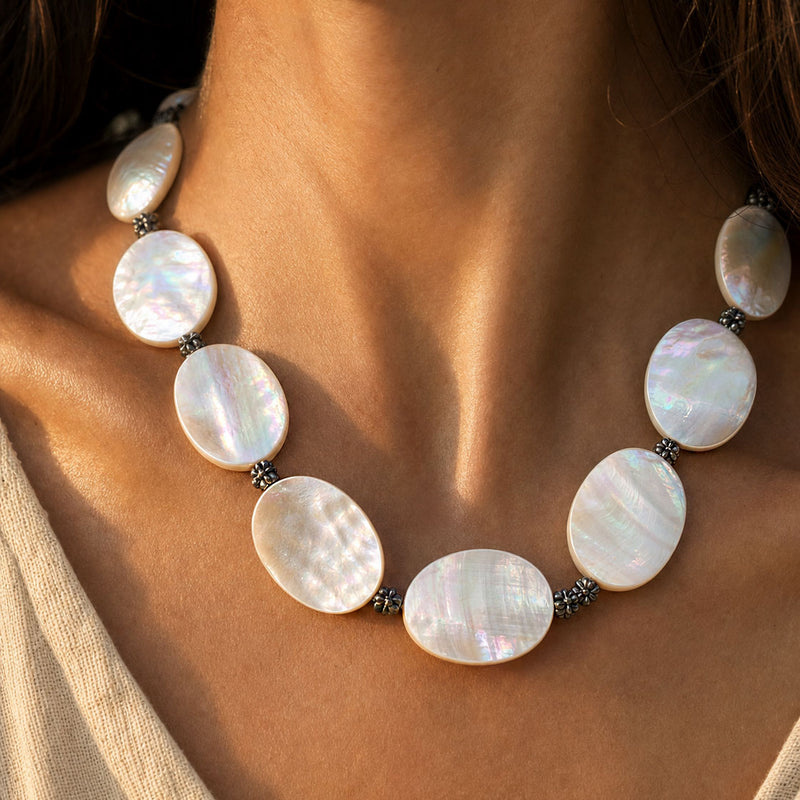 A close-up of a woman's neck and upper chest wearing a necklace made of large oval iridescent shell mother of pearl beads separated by small dark metallic spacer beads.