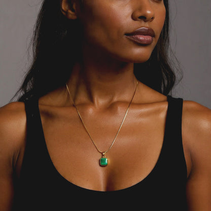 Woman wearing a black tank top and gold necklace with a green pendant against a gray background