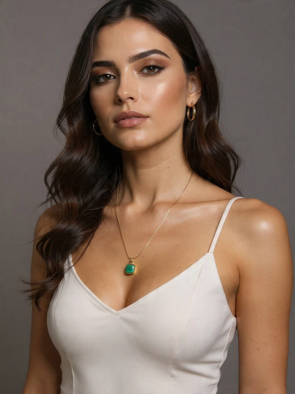 Woman wearing a white top and gold necklace with a green pendant against a gray background