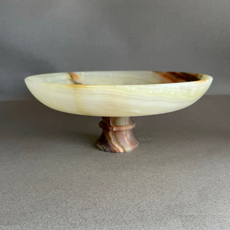 Decorative stone bowl with a pedestal base on a gray background Hand-carved cream onyx pedestal bowl with honey and blush veining, styled as a sculptural fruit bowl on a neutral background
