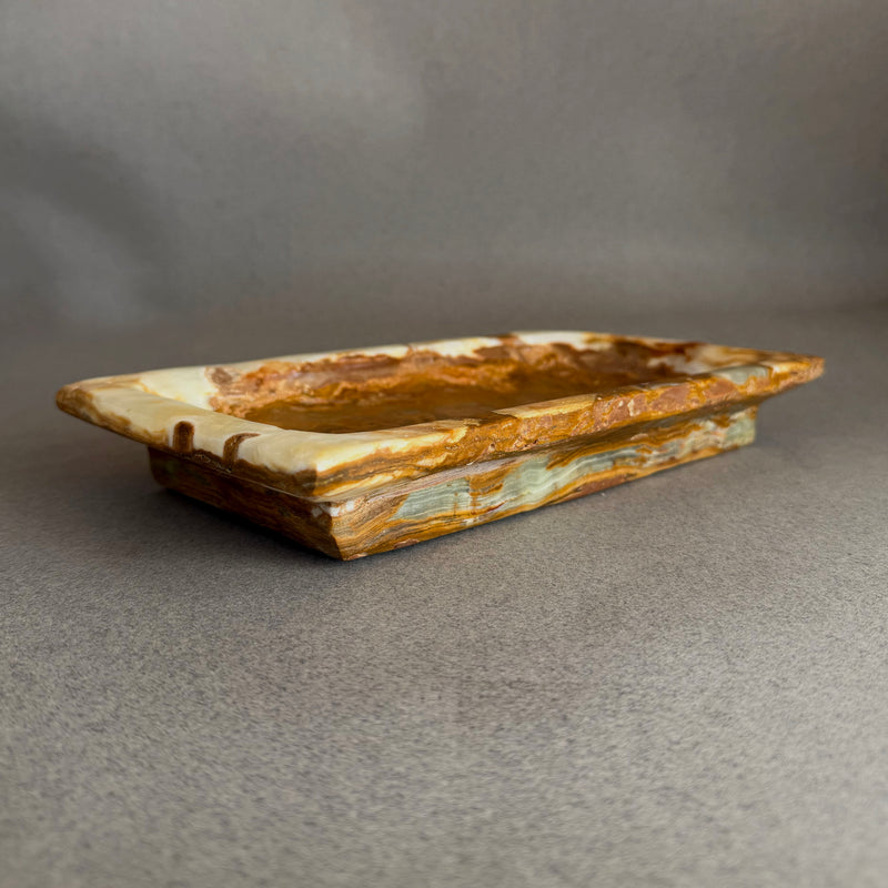 Hand-carved raw onyx vanity tray in cream, honey, and warm brown tones styled with perfume, watch, and hand cream on a neutral surface
