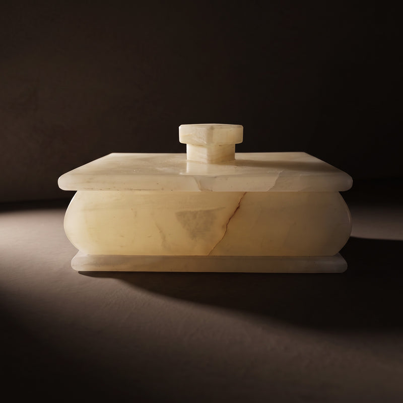 Cream and blush onyx keepsake box with lid seen straight on, its pale translucent body and stepped base lit by a low beam of light against a dark backdrop, with a square stone knob on the lid