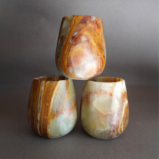 Three hand-carved saffron and sienna onyx curved vessels stacked together, warm amber and burnt orange stone veining on cream, grey studio surface