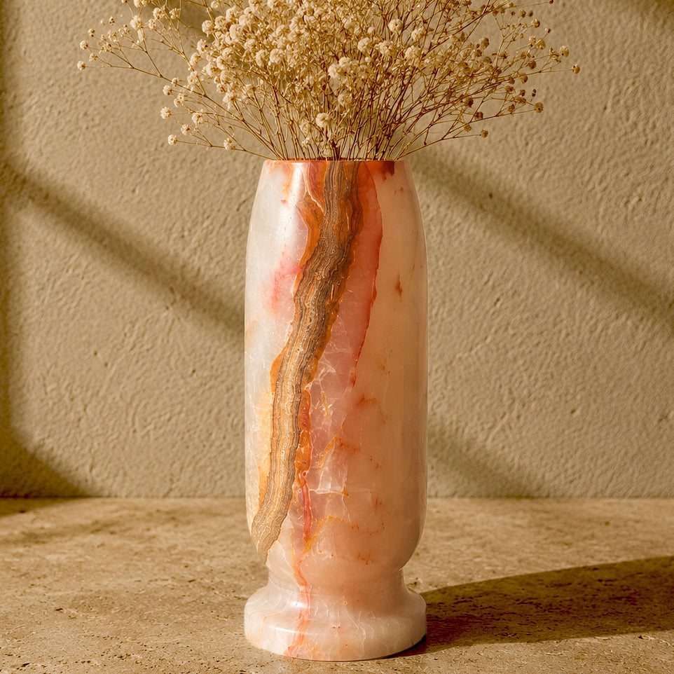 A tall cylindrical vase made of pink and cream marbled stone with a vertical brown veining, filled with dried baby's breath flowers, set on a textured beige surface against a matching beige wall with window shadow patterns.