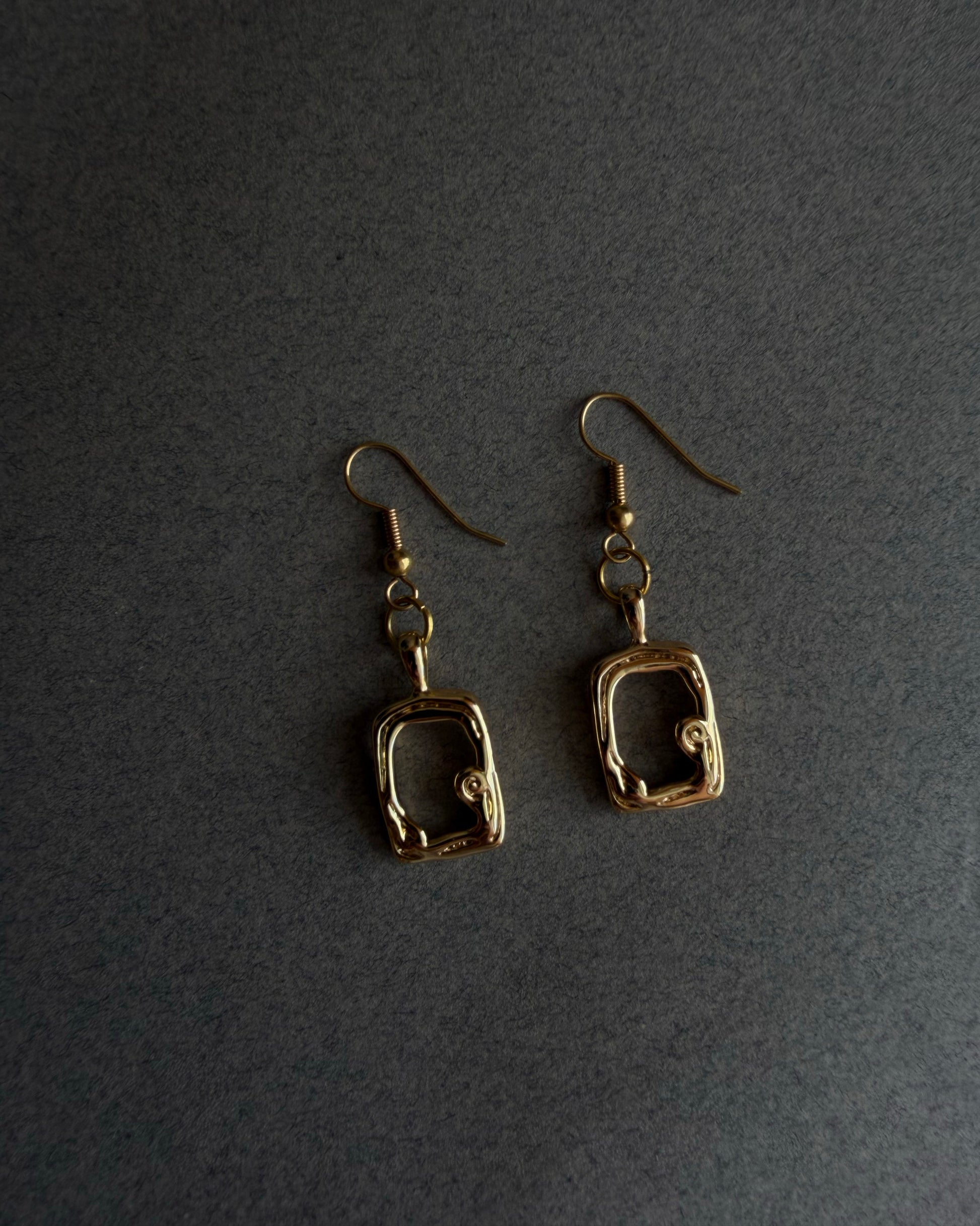 Pair of gold square-shaped earrings on a dark gray backgroundPair of handmade gold sculptural drop earrings on neutral fabric background, modern minimalist statement jewelry
