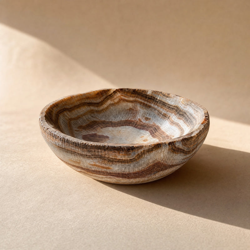Onyx stone bowl styled on a wood table with pinecones, showing rust and cocoa striations in warm light.
