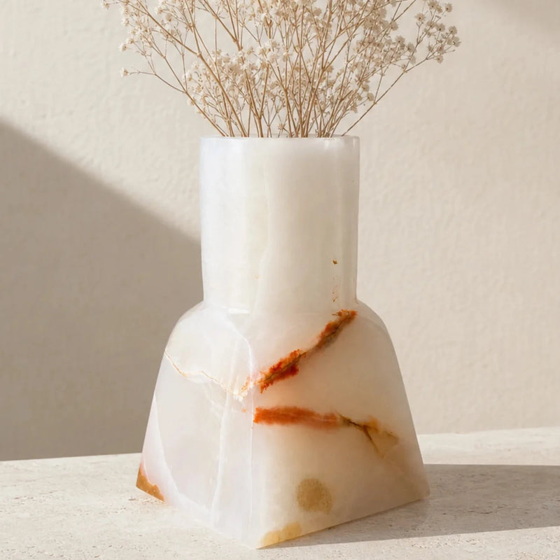 A white onyx vase with natural orange-brown veining, holding dried baby's breath flowers, set on a light stone surface against a warm beige wall with soft diagonal shadows.