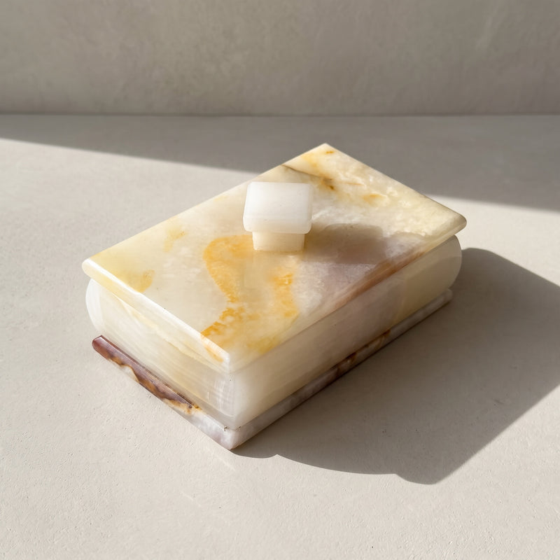 Ivory and honey onyx ring and trinket box with lid, seen from above at an angle: a creamy translucent lid swept with golden-honey veining and a blush patch, a white square stone knob and a stepped base edged in rust, on pale plaster in a slant of sunlight